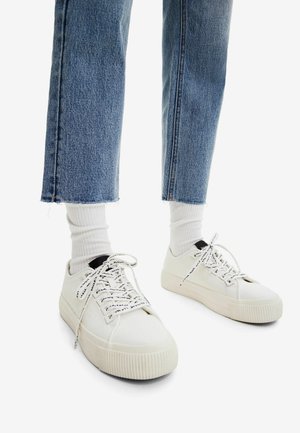 White canvas sneakers with rubber soles, featuring patterned laces and a black accent on the heel, paired with blue cropped jeans and white socks.