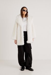 Λευκό παλτό από ψεύτικη γούνα με φαρδύ γιακά, φορεμένο πάνω από ένα ανοιχτόχρωμο πουκάμισο και μαύρο παντελόνι cargo. Το μοντέλο φοράει μαύρα γυαλιά ηλίου και μπότες με κορδόνια.