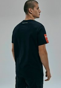 Czarny bawełniany t-shirt z czerwoną łatą na lewym rękawie i subtelnym białym brandingiem na tylnej części kołnierzyka. Krótkie rękawy, regularny krój.