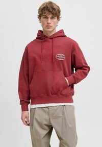 Sweat à capuche en coton rouge avec une poche frontale, un cordon de serrage pour la capuche et une écusson logo sur la poitrine, associé à un pantalon beige ample.