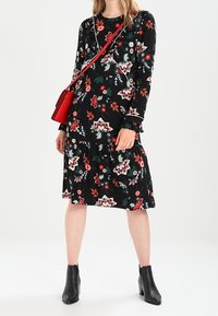Černé květinové šaty s dlouhým rukávem, které mají červené a zelené akcenty, s tyrkysovým designem, doplněné červenou crossbody kabelkou a černými kotníkovými botami.