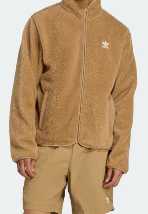 Man draagt een beige sherpa fleece jas met rits en bijpassende beige korte broek, beide met een wit Adidas-logo aan de linkerkant.