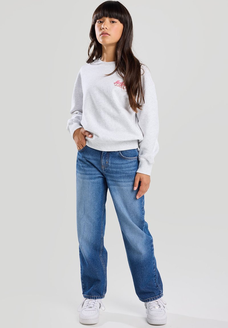 Harmaa huppari, jossa on pieni punainen logo, yhdistettynä sinisiin suoriin farkkuihin ja valkoisiin tennareihin, korostaen rentoa nykyaikaista tyyliä.