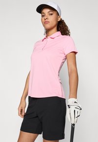 Ružová polokošeľa s golierom, krátkymi rukávmi a priliehavým strihom; v kombinácii s čiernymi šortkami a bielou čiapkou. Drží golfovú palicu.