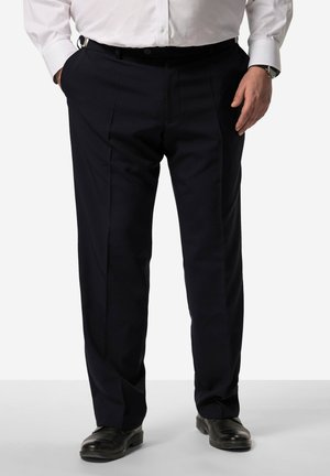 Mand iført sorte bukser, sorte lædersko og en hvid skjorte med knapper, stående med hænderne delvist i lommerne.