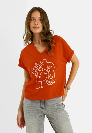 Femme aux cheveux ondulés portant un T-shirt orange à col en V avec un visage en dessin linéaire blanc et un jean gris, debout avec une main sur la hanche.