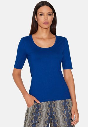 Blaue schulterfreie Bluse mit Rundhalsausschnitt, gefertigt aus einem glatten, dehnbaren Stoff. Das Model trägt gemusterte Hosen mit geometrischen Details.