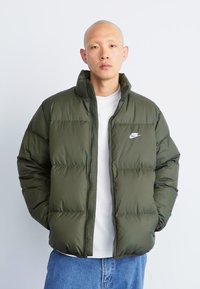 Man wearing olive green Nike puffer jacket over white shirt and blue jeans, hands in pockets, standing against plain background.