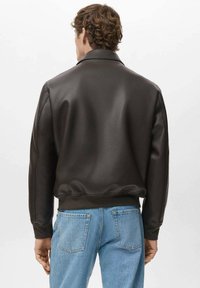 Chaqueta marrón de cuero sintético con una textura suave, puños y cinturilla acanalados; combinada con vaqueros denim azul claro. Vista trasera.