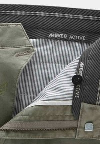 Olivenfarvet stof med en tekstureret overflade, stribet indvendig foring, sorte accenter og synlige sømme. Har lynlås og knaplukning.