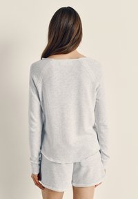 Pruunide pikkade juustega naine, kes kannab helehallit pika varrukaga topi ja sobivaid lühikesi pükse, seisab palja beežika seina ees.