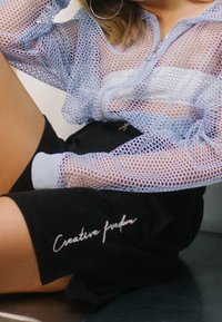 Osoba w jasnofioletowej siatkowej kurtce na białej bluzce, czarnych szortach z napisem "Kreatywna wolność", siedząca z nogami ugiętymi.