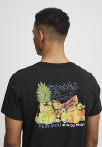 Juodas medvilnės marškinėliai su spalvingu vaisių grafiniu atvaizdu ant nugaros, pažymėti "Florida’s Finest" tekstu ir tekstūruotu dizainu.