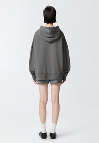 Sudadera oversized gris oscuro con capucha, con puños acanalados, llevada sobre pantalones cortos de cuadros. El modelo lleva calcetines blancos y calzado negro. Fondo liso.