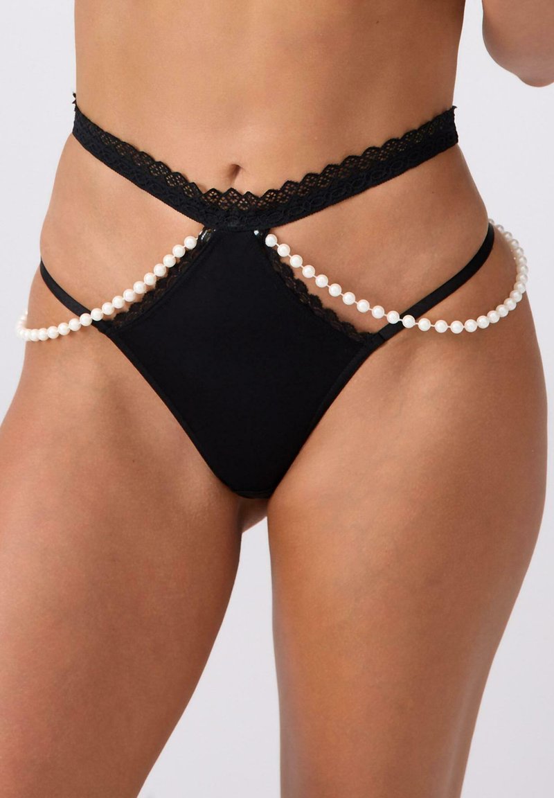 Czarny koronkowy dół bielizny z perłowymi detalami noszony na biodrach i górnej części ud kobiety, na jednolitym jasnym tle.