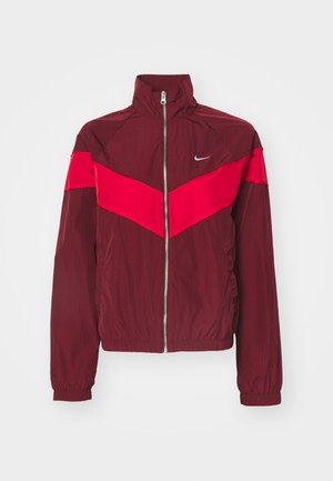 Bordo zip-up jakna z visokim ovratnikom, ki ima širok rdeč trak čez prsni del in ramena, elastične manšete in majhen logotip.