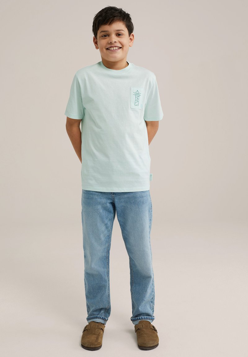 Gaiši zilā kokvilnas T-kreklā ar īsām piedurknēm, uz kreisā krūšu kabatas ir bākas grafika. Valkāts ar zilām džinsām un brūnām kurpēm.