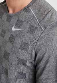 Šedé sportovní tričko s texturovaným kostkovaným vzorem, bílým logem Nike a jemnými šedými pruhy na rameni.