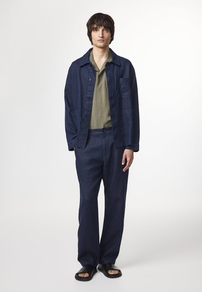 Giacca in lino blu scuro con vestibilità rilassata e tasche frontali, abbinata a pantaloni coordinati e una camicia con colletto verde chiaro. Sandali neri.