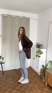 Brown leather oversized jacket, white t-shirt, light grey skinny jeans, and white sneakers. Natural wood floor and indoor plants in background.