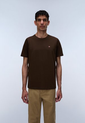 Homme debout face à l’avant, portant un t-shirt marron à manches courtes avec un petit logo sur la poitrine et un pantalon beige sur un fond clair uni.