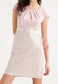 Robe à manches courtes avec un haut rose clair, un col plissé et une jupe crème à motifs géométriques. Texture lisse dans l'ensemble.