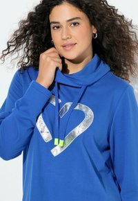 Modrá mikina s kapucí s límcem a kovovým stříbrným srdcem jako grafikou. Má modré šňůrky s neonově zelenými konci. Měkká textilie.