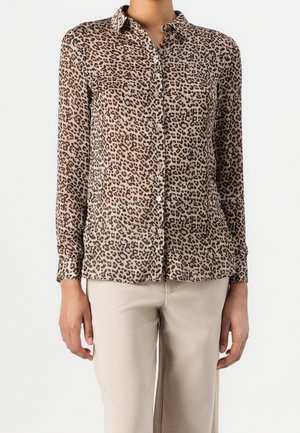 Femme portant une chemise à manches longues imprimé léopard avec boutons, associée à un pantalon beige clair sur fond blanc uni.