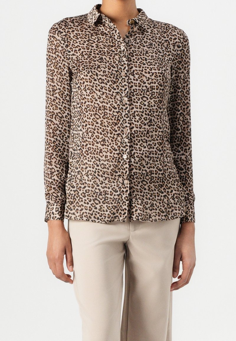 Femme portant une chemise à manches longues imprimé léopard avec boutons, associée à un pantalon beige clair sur fond blanc uni.