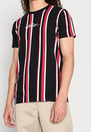 Person iført en sort kortærmet skjorte med lodrette røde, hvide og sorte striber og beige bukser, stående op ad en ensfarvet væg.