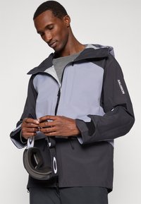 Homem a ajustar uma jaqueta Salomon preta e cinza, segurando um par de óculos de ski pretos com o logótipo branco da Oakley, contra um fundo simples.