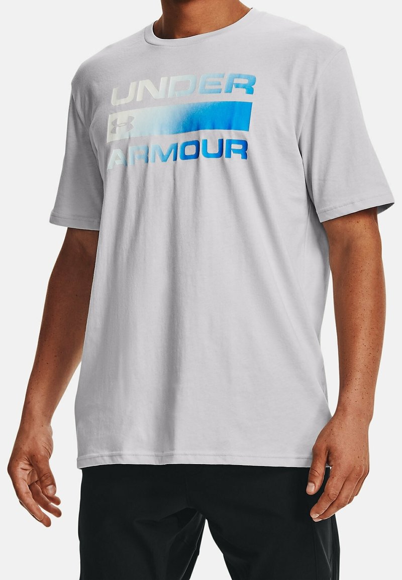 Camiseta de algodón blanca con texto bold "UNDER ARMOUR" en tonos azules en degradado. Mangas cortas, ajuste regular, textura suave y diseño de cuello redondo.