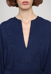 Kobieta ubrana w granatowy strój z głębokim dekoltem w kształcie litery V oraz wyraźnymi przeszyciami na klatce piersiowej i ramionach.