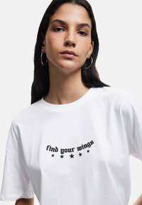 Witte katoenen T-shirt met de tekst "find your wings" in zwarte schrift en kleine zwarte steraccenten, met een losse, comfortabele pasvorm.