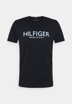 Schwarzes T-Shirt mit kurzen Ärmeln und Rundhalsausschnitt mit dem Schriftzug „HILFIGER MCMLXXXV“ in großen, reflektierenden hellblauen Buchstaben über der Brust.