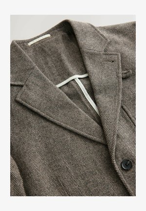 Brūns herringbone blazeris ar vīlēm, vienu pogu slēgšanu, teksturētu audumu un kontrastējošu iekšējo oderi. Redzama melna poga.