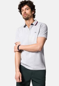 Jasnoszara koszulka polo z granatowymi akcentami przy kołnierzyku i czerwoną paskiem, krótki rękaw, z wyszytym logo. Noszona na ciemnozielonych spodniach.