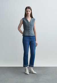 Femeie purtând un top gri fără mâneci cu nasturi, blugi scurți albaștri cu ornamente florale, șosete albe și pantofi crem cu toc jos.