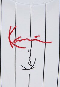 Bílá tkanina s tenkými vertikálními černými pruhy a červeným výšivkovým designem ve stylu podpisu napříč středem.