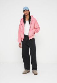 Ružová oversized zipová mikina, biele tričko, čierne široké nohavice a svetlomodrá čiapka. Hnedé čižmy dopĺňajú uvoľnený outfit.