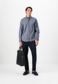 Hombre con una camisa de botón azul con estampado y pantalones oscuros, sosteniendo una bolsa tote de cuero negra, de pie frente a un fondo blanco.