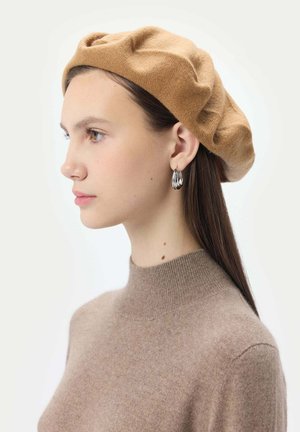 Beret en tricot marron avec un design plissé, associé à des boucles d'oreilles en argent pendantes. Pull doux de couleur beige clair avec un col montant et une texture fine.