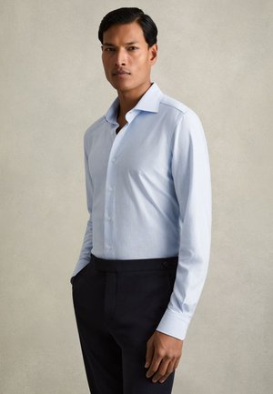 Mies, jolla on lyhyet tummat hiukset, pukeutuneena vaaleansiniseen raidalliseen kauluspaitaan ja mustiin housuihin, seisoo yksivärisen beigen taustan edessä.