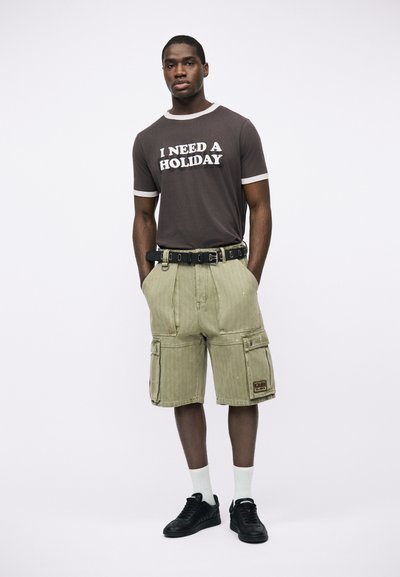 Άνδρας που φοράει μαύρο μπλουζάκι με την επιγραφή "ΧΡΕΙΑΖΟΜΑΙ ΔΙΑΚΟΠΕΣ", μπεζ cargo σορτς με μαύρη ζώνη, λευκές κάλτσες και μαύρα αθλητικά παπούτσια, στέκεται με τα χέρια στις τσέπες.