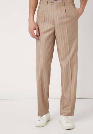Hombre con pantalones beige de rayas finas y zapatillas blancas, de pie con una mano en el bolsillo y la otra relajada al lado.