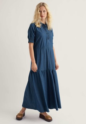 Femeie cu păr blond ondulat, purtând o rochie midi albastră cu guler volanat și mâneci bufante, stând în sandale maro cu platformă, pe un fundal simplu.