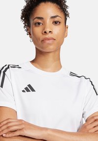 T-shirt sportivo bianco realizzato in tessuto leggero, con strisce nere sulle spalle e logo. Il design è a girocollo con una vestibilità rilassata.