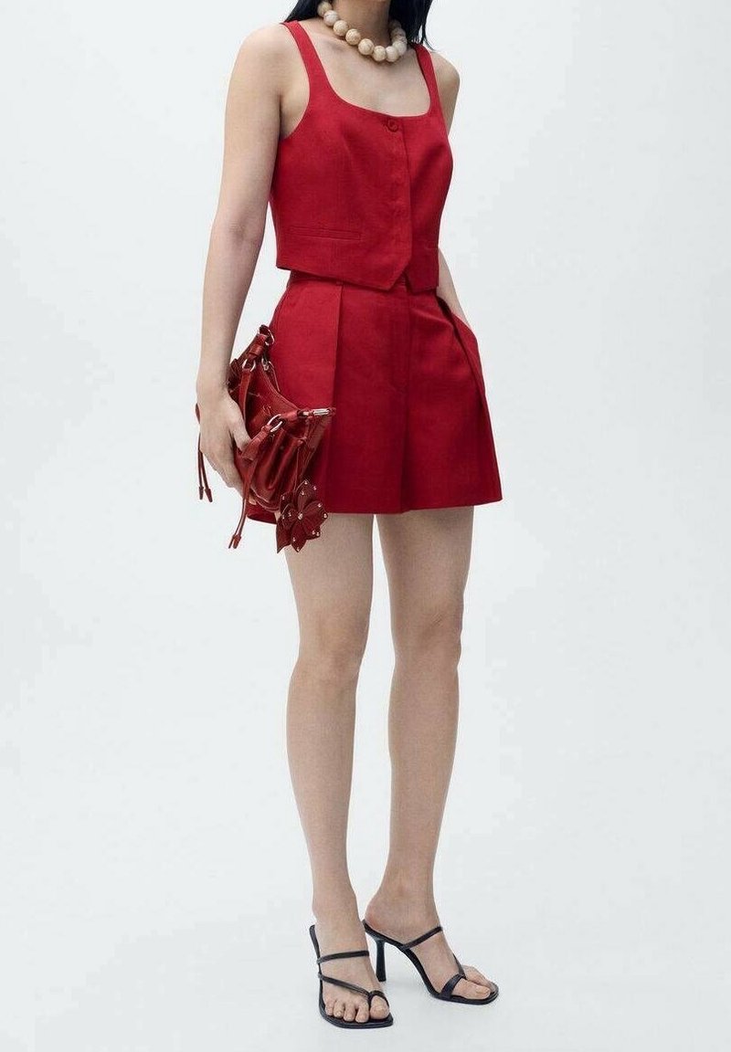 Femme portant un haut rouge sans manches à boutons et une jupe assortie, tenant un sac à main rouge, accessoirisée d'un grand collier de perles et de sandales noires à talons.
