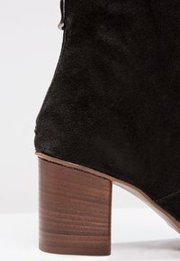 Bottine en daim noir avec un bout rond et un talon en bois empilé. Dispose d'une fermeture éclair sur le côté. Texture lisse.