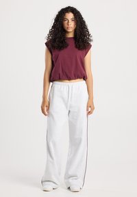 Jeune femme avec des cheveux bouclés portant un haut sans manches bordeaux et un pantalon de survêtement blanc ample se tenant devant un fond blanc uni.